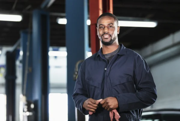 A smiling mechanic in navy coveralls holding a shop rag inside an auto repair shop service bay.