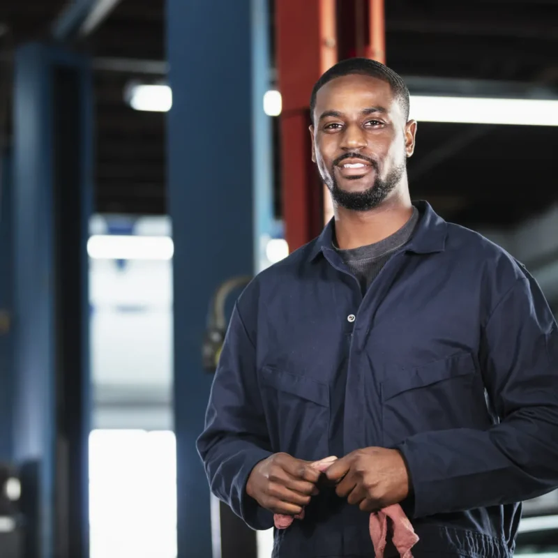 A smiling mechanic in navy coveralls holding a shop rag inside an auto repair shop service bay.