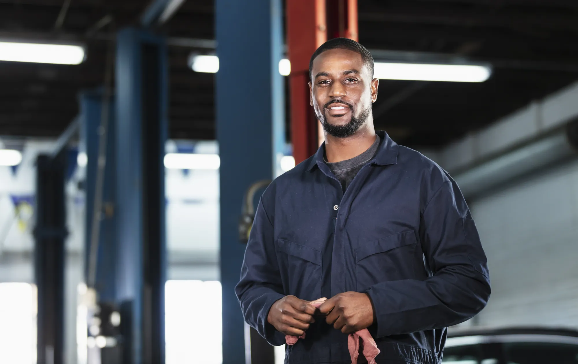 A smiling mechanic in navy coveralls holding a shop rag inside an auto repair shop service bay.