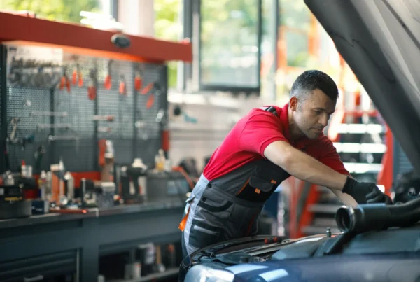 A mechanic in a red shirt and work overalls leans under the open hood of a car inside a repair shop.
