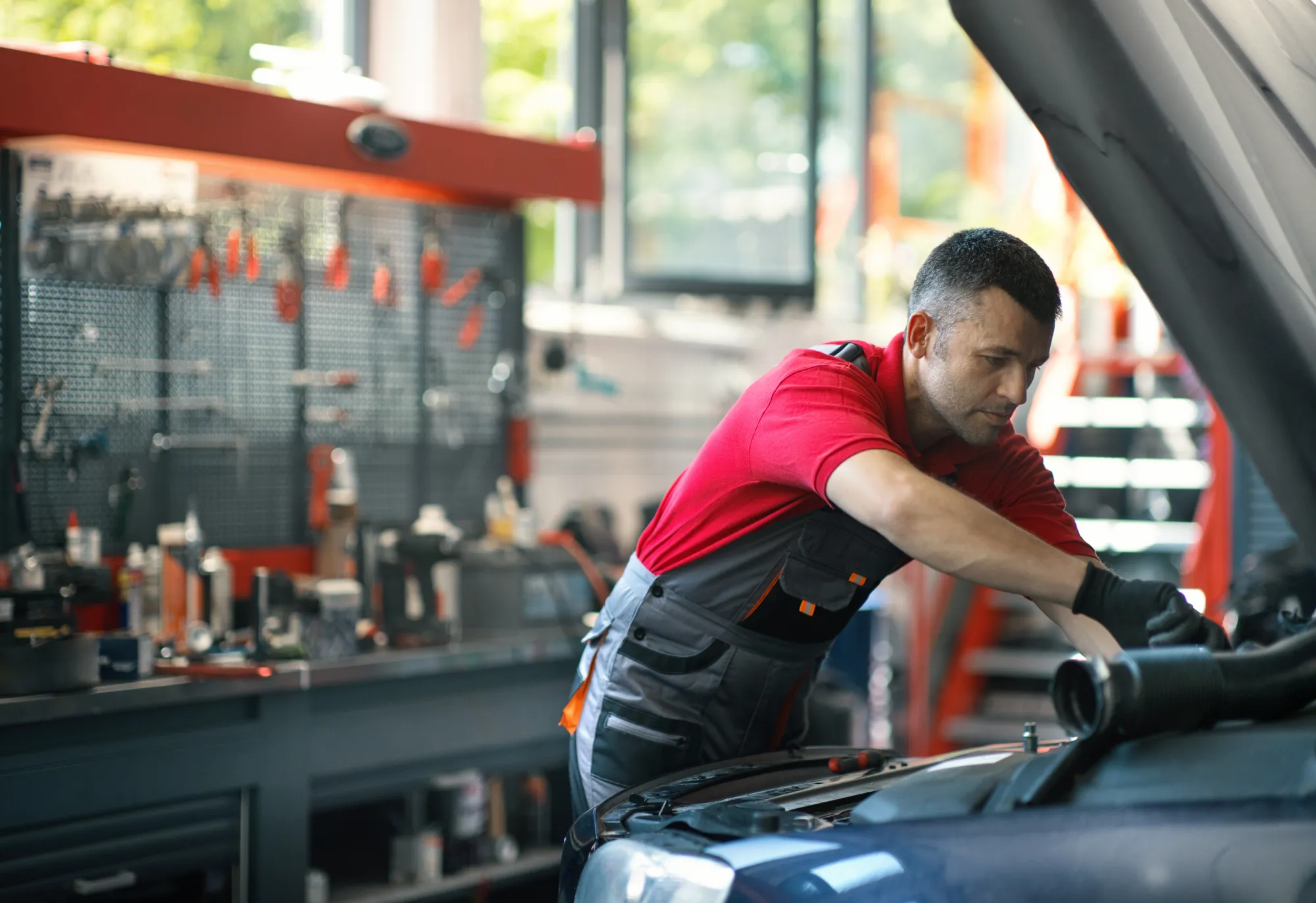 A mechanic in a red shirt and work overalls leans under the open hood of a car inside a repair shop.
